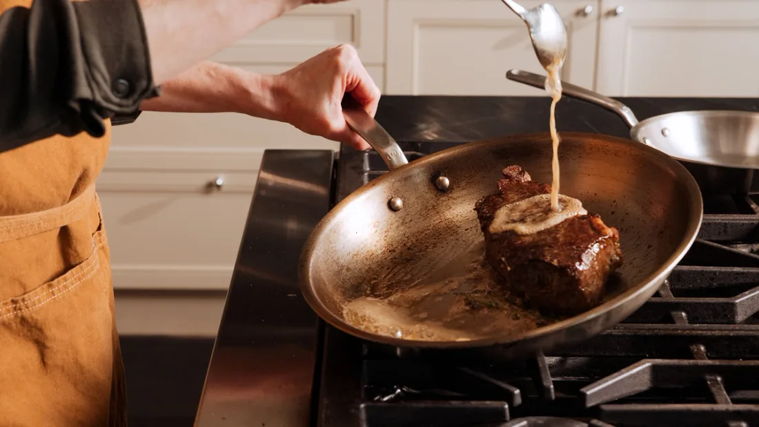 A cook spoon-basting a steak with foaming butter in a stainless steel frying pan on a gas range