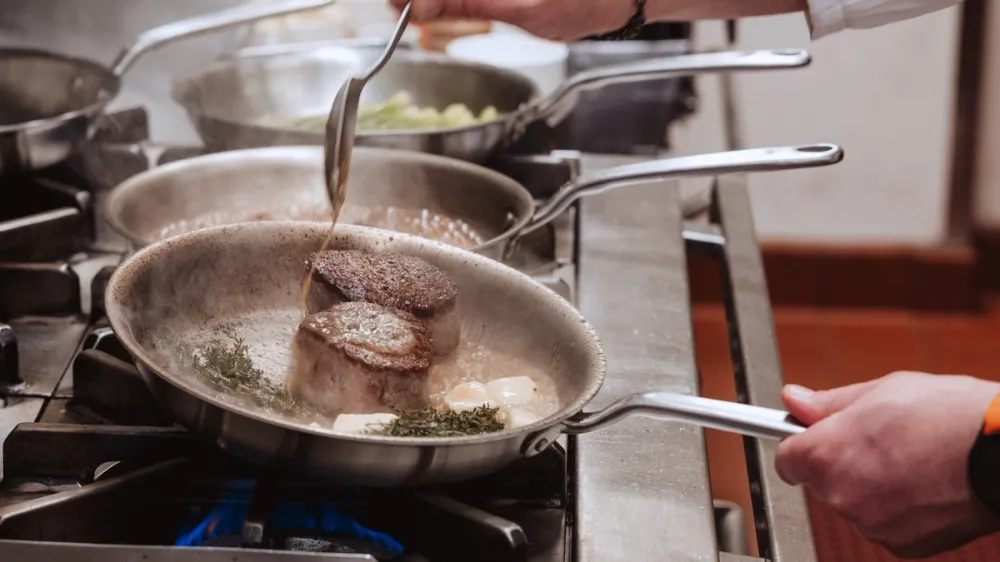 A cook basting a thick cut of meat with a spoon on a restaurant range, with several stainless steel pans working at once