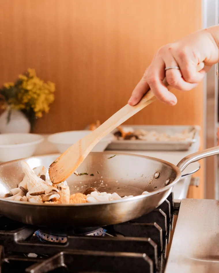 A hand tossing mushrooms and shallots in a stainless steel frying pan over a flame