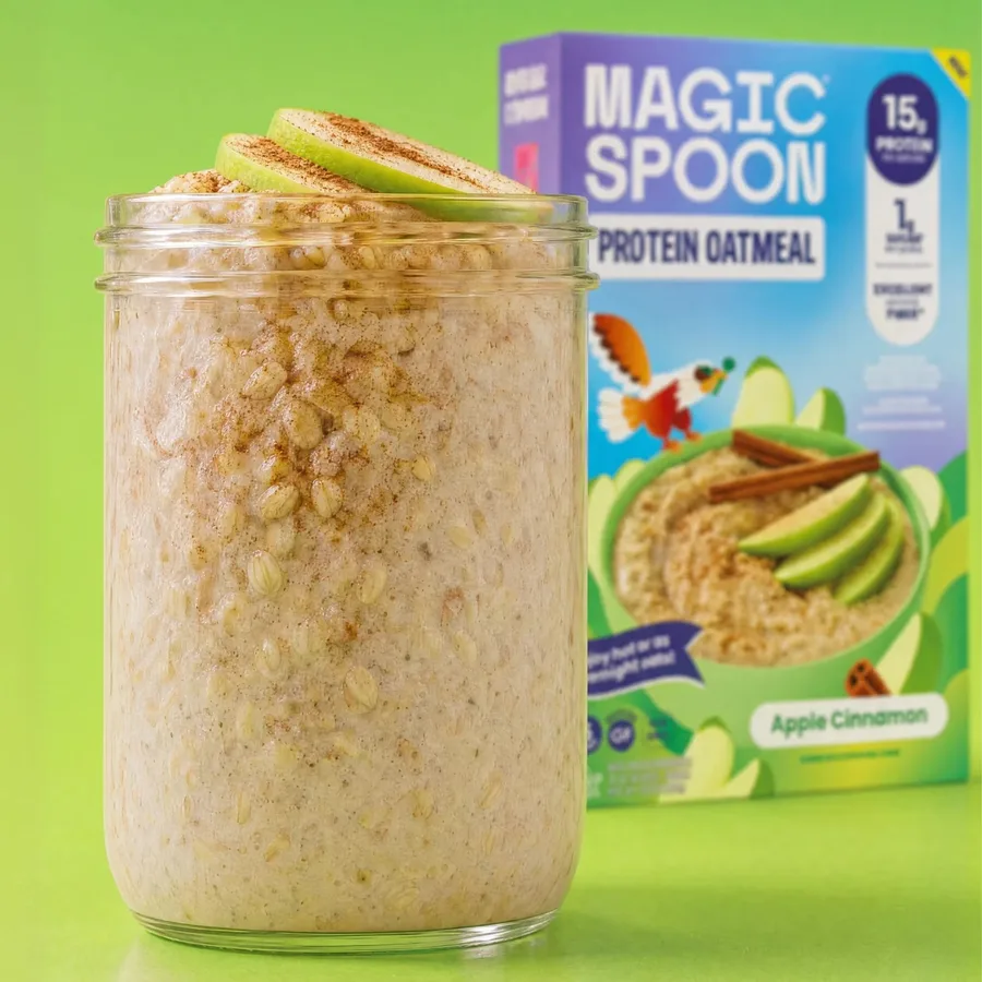 A glass jar of Apple Cinnamon overnight oats topped with apple slices and cinnamon, in front of the Magic Spoon Protein Oatmeal box