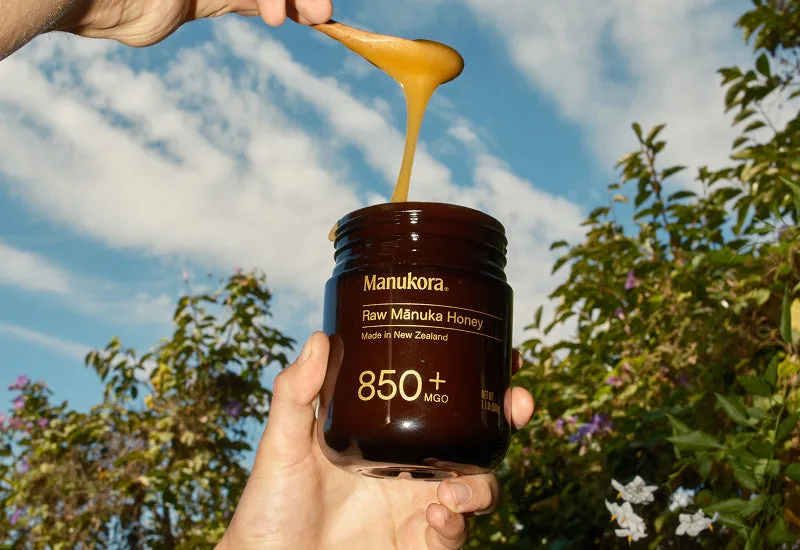 Hand holding an amber jar of Manukora Raw Manuka Honey 850 plus MGO with honey dripping from a spoon