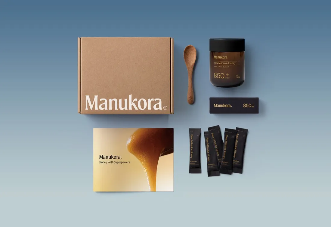 Manukora starter kit laid out flat: shipping box, wooden spoon, amber 850 plus jar, guidebook and honey sticks