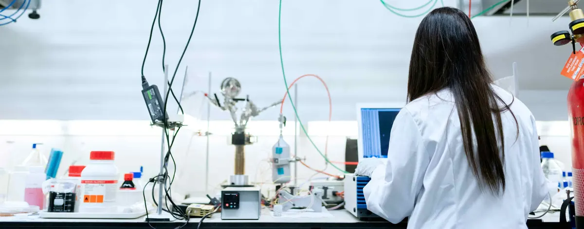 Wide view of an analytical laboratory bench with glassware, coloured tubing, reagent bottles and sample racks, an analyst in a white coat working at a screen on the right
