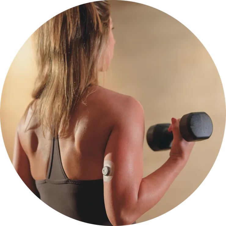 A woman lifting a dumbbell with a small round glucose sensor visible on the back of her arm.