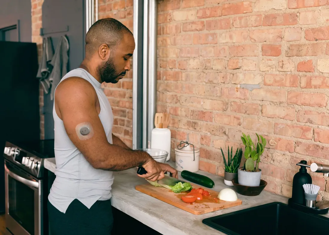 A man wearing a glucose sensor on his upper arm chopping vegetables at a kitchen counter.
