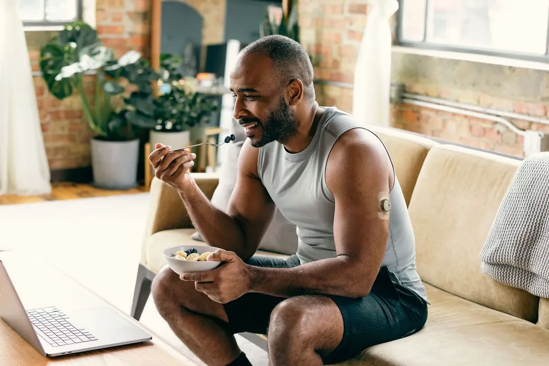 A man eating a bowl of food at home, a glucose sensor visible on the back of his arm.