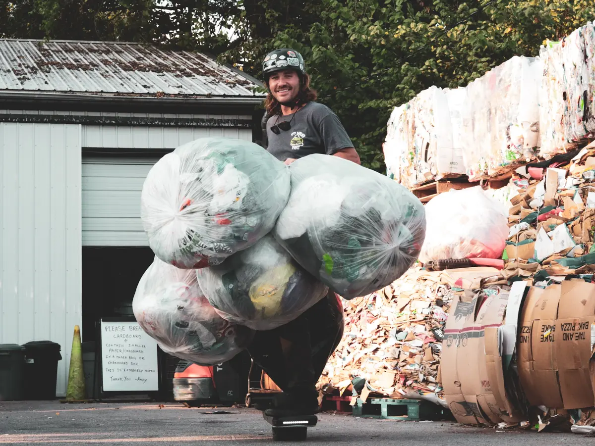 A rider carries several large bagged loads on a Onewheel in front of a wall of baled recycling.
