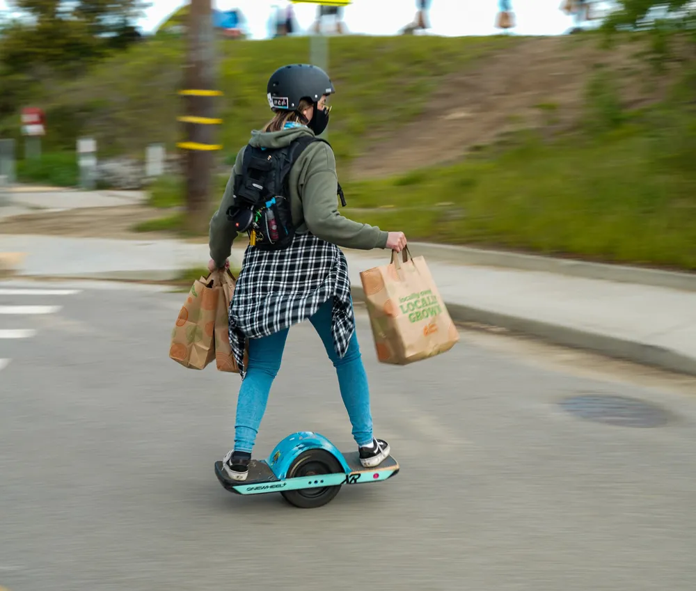 A rider carries two paper grocery bags while riding a Onewheel down a street.