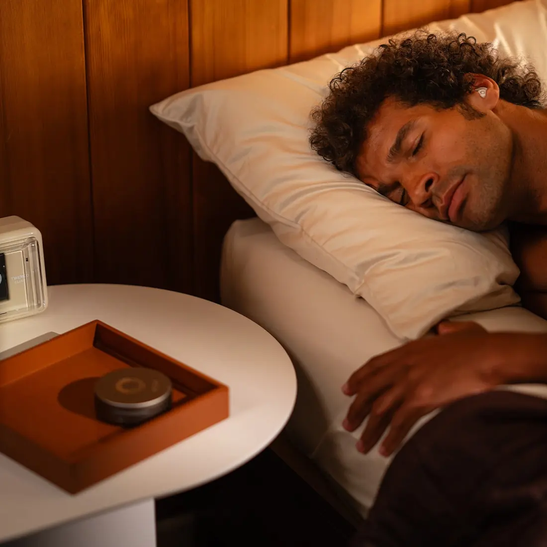 A man asleep in bed at night with a lamp and the charging case on the bedside table
