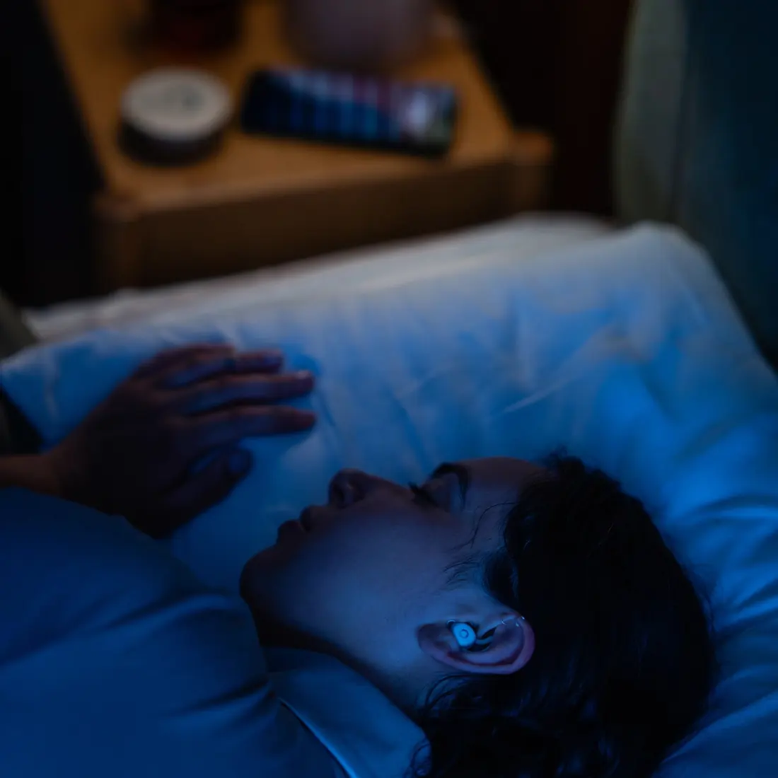 A man asleep in bed at night wearing sleep earbuds, with the charging case glowing on the bedside table