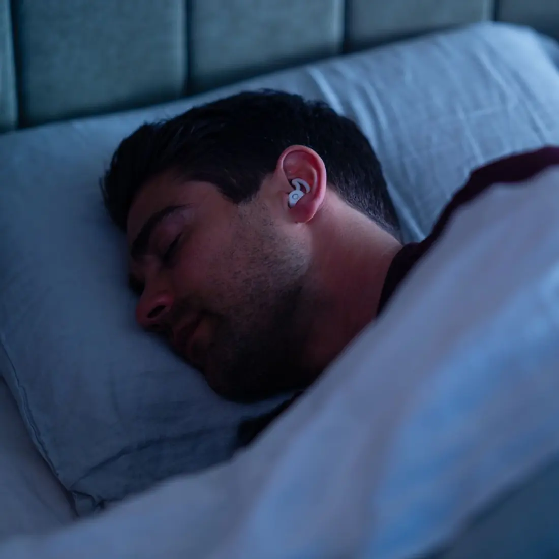 A man asleep on his side wearing a sleep earbud, lit by a soft blue night light