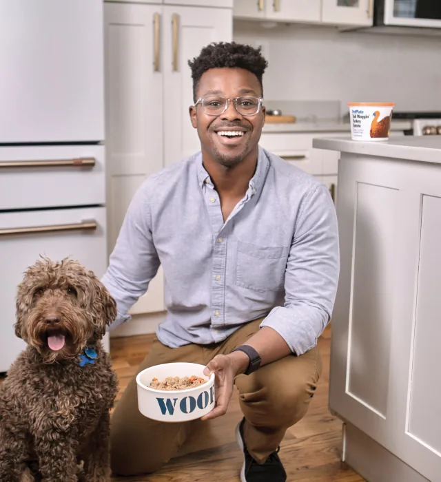 PetPlate founder Renaldo Webb kneeling in a kitchen beside a dog and a full bowl