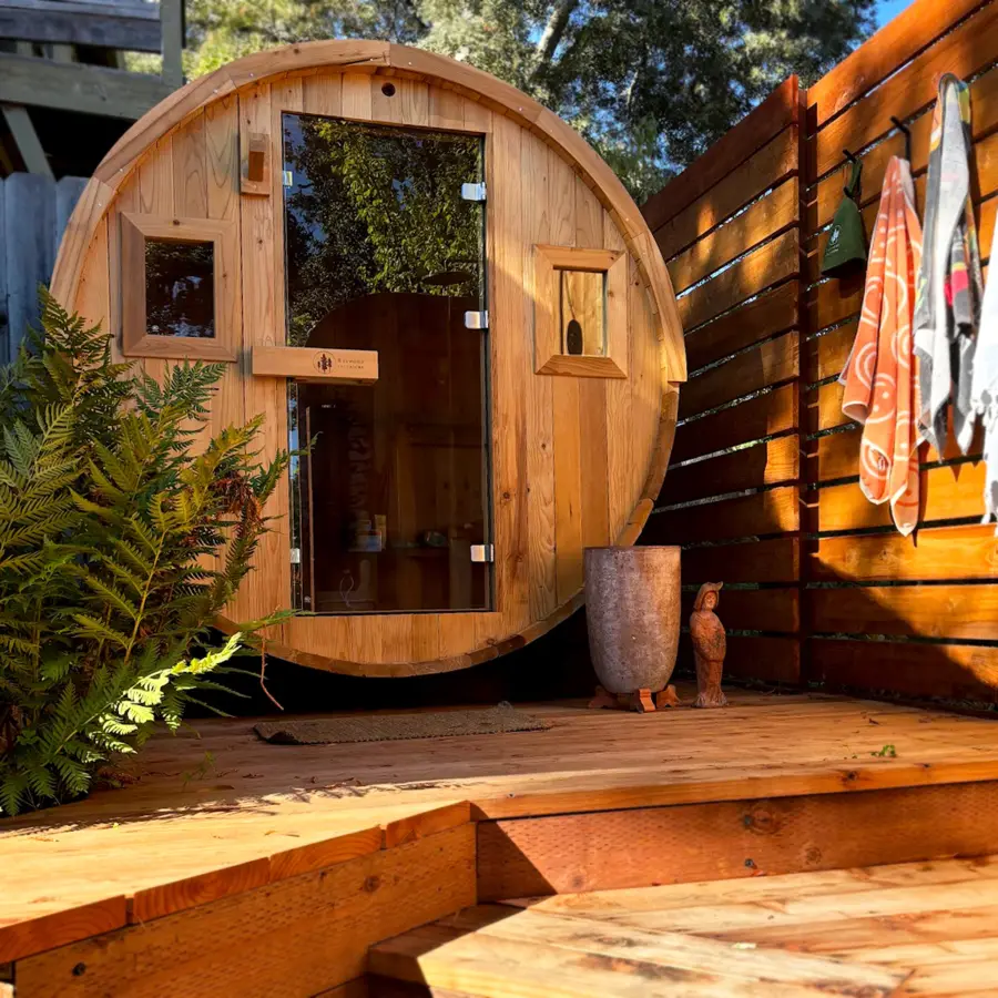Six person barrel sauna installed on a paved patio beside a wooden fence, with a towel rail and mat outside the door