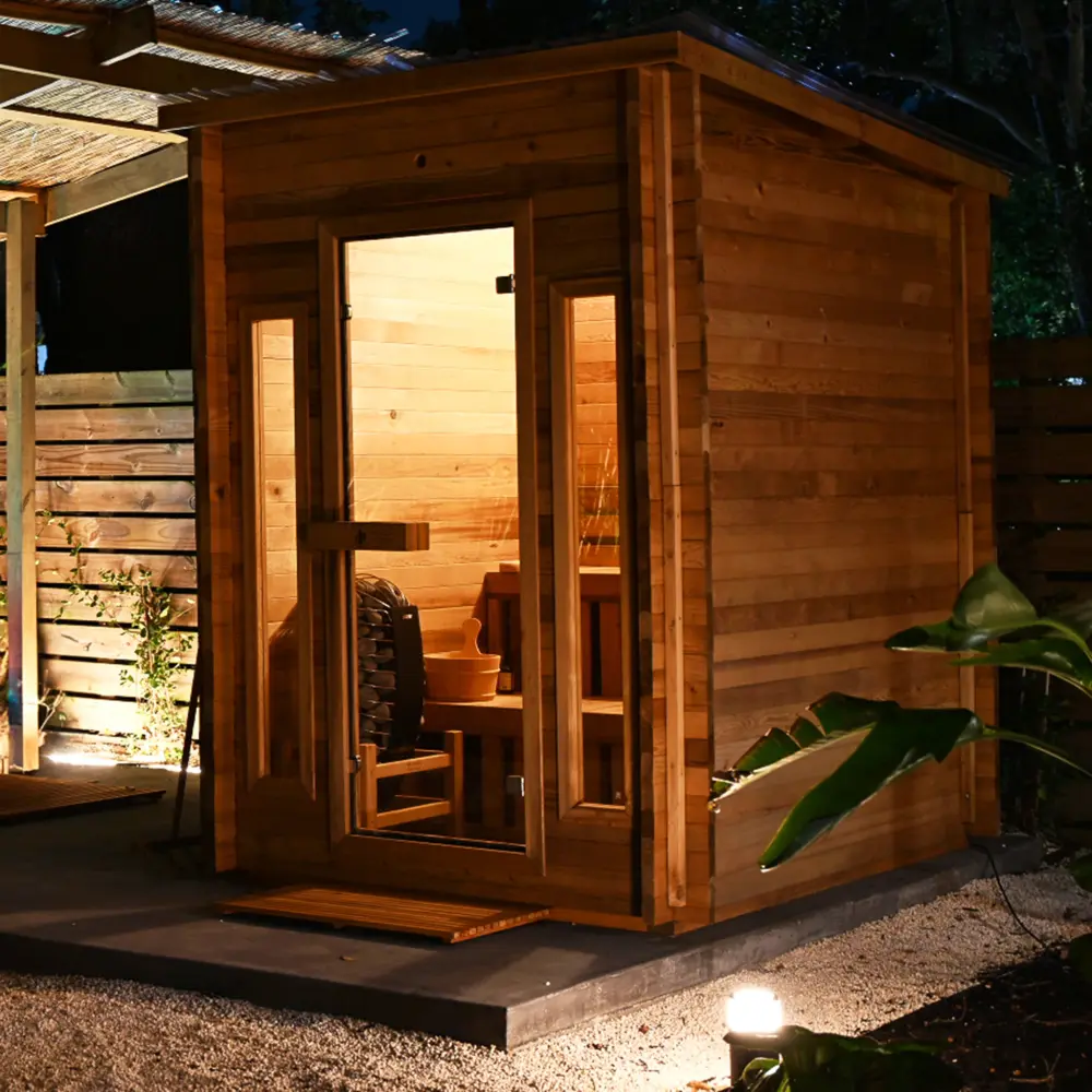 Cabin outdoor sauna installed on a dark raised platform set into a gravel bed, with landscape lighting at the base