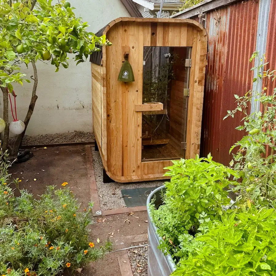 A two person outdoor sauna installed in a narrow side yard on a gravel and paver base, between a stucco wall and a corrugated metal fence