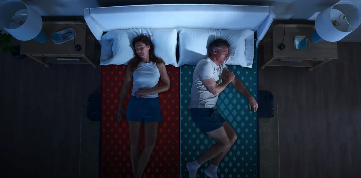 Two sleepers seen from above on one bed, the left half of the mattress shown in warm red and the right half in cool blue