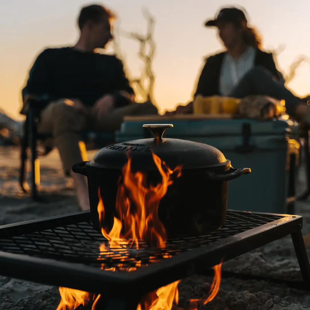 A Smithey cast iron Dutch oven cooking over an open campfire at dusk with two people seated behind it.