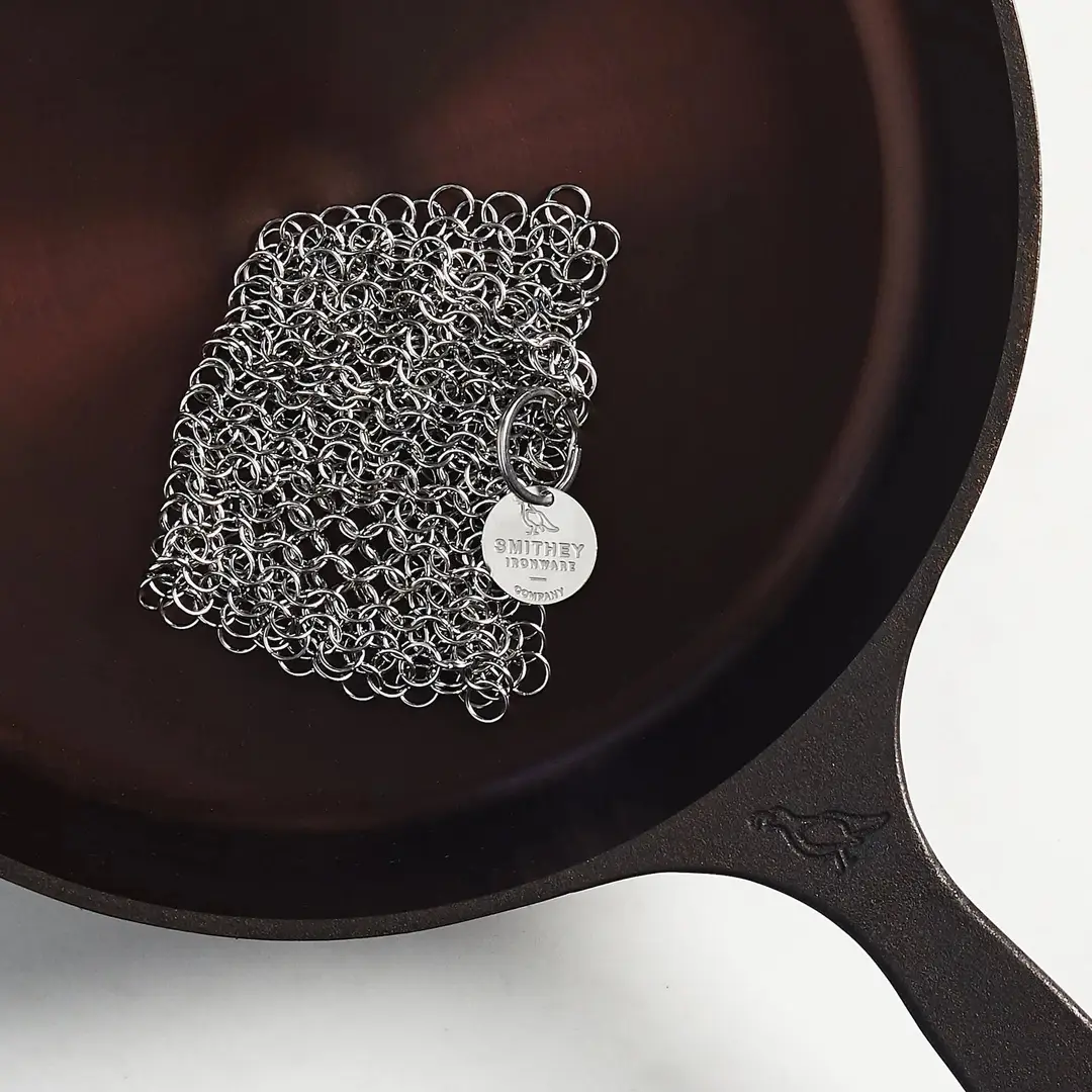 A stainless steel chainmail scrubber resting on the polished interior of a Smithey skillet.