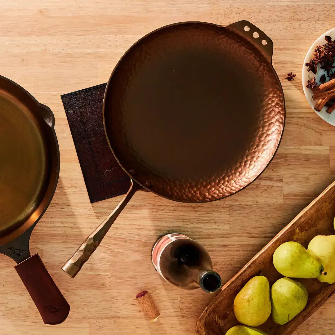 A hand forged Smithey carbon steel farmhouse skillet with a leather handle sleeve on a wooden counter.