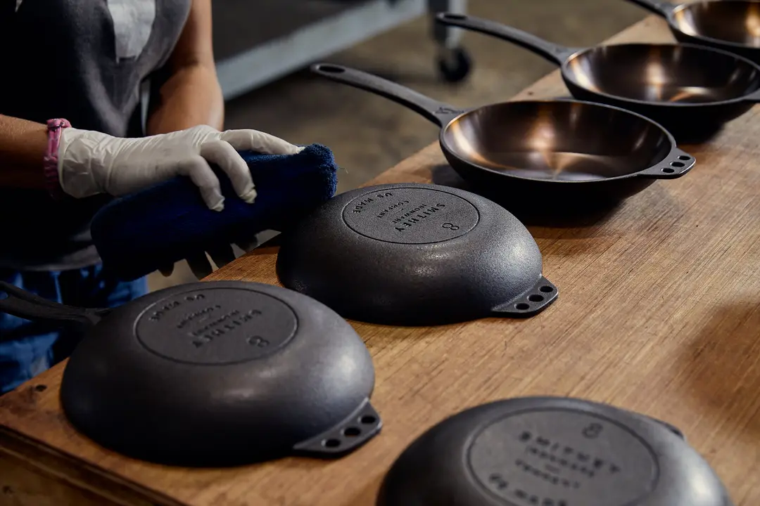 A worker in gloves handling raw and finished Smithey skillets on a wooden workbench.