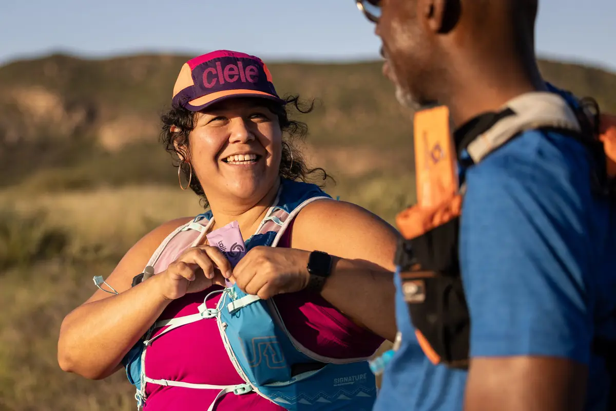 Two trail runners in vests on an open ridge, one adjusting a soft flask, a single serving packet visible in the other runner's chest pocket