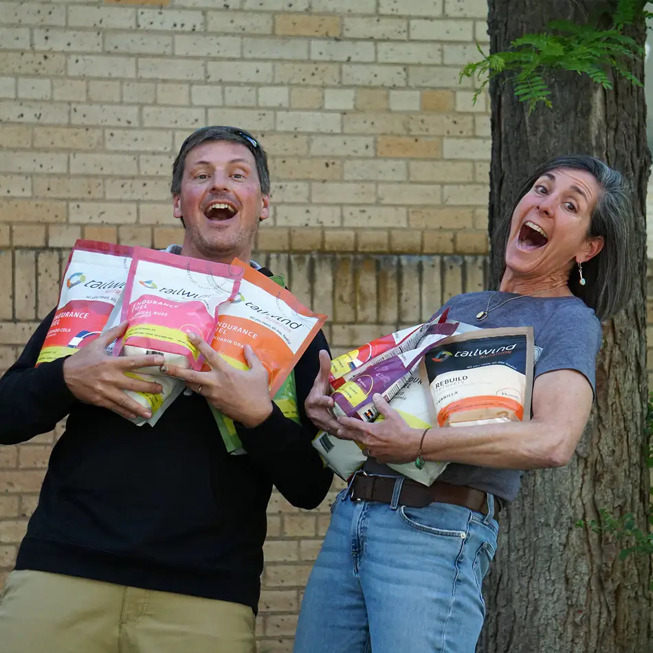 Two people outside a brick building in Durango, arms full of Tailwind Endurance Fuel bags, laughing