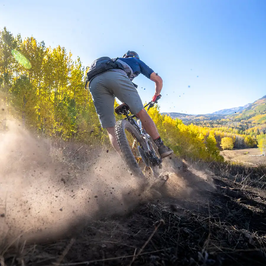 Mountain biker throwing up a wall of dust on a dry Colorado trail with aspens turning yellow behind