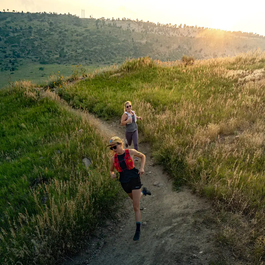 Two runners on singletrack crossing a high meadow in golden evening light