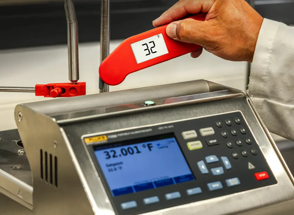 A hand in a white lab coat holding a red Thermapen reading 32 next to a benchtop reference instrument displaying 32.001 degrees Fahrenheit