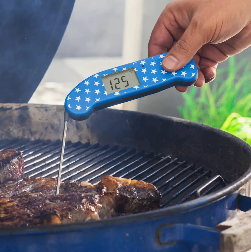 A hand holding a star-spangled Thermapen ONE reading 125 degrees, probing a steak over a charcoal kettle grill