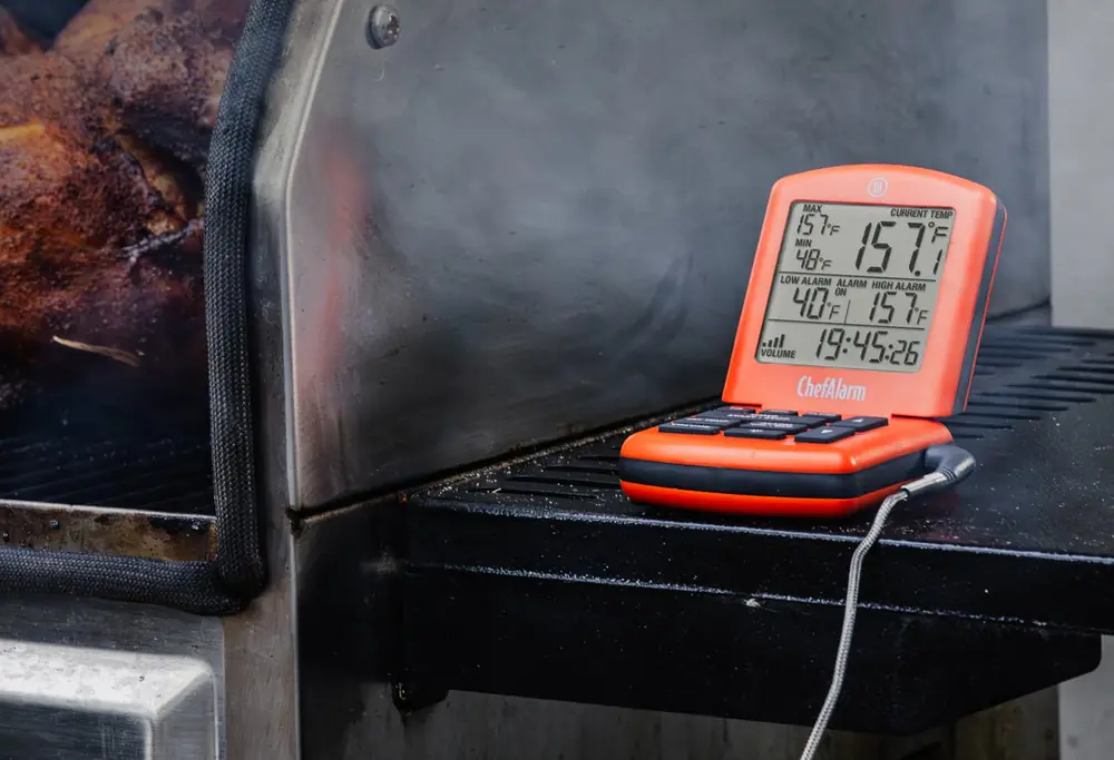 An orange ChefAlarm alarm thermometer sitting directly on the grate of a smoker beside cooking meat, smoke drifting across the frame