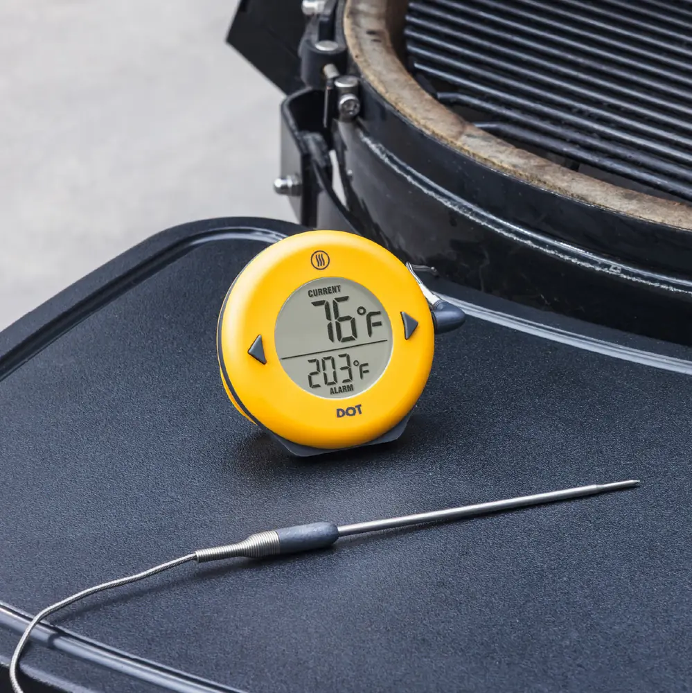 A yellow DOT alarm thermometer with its probe coiled, sitting on the side table of a ceramic kamado grill