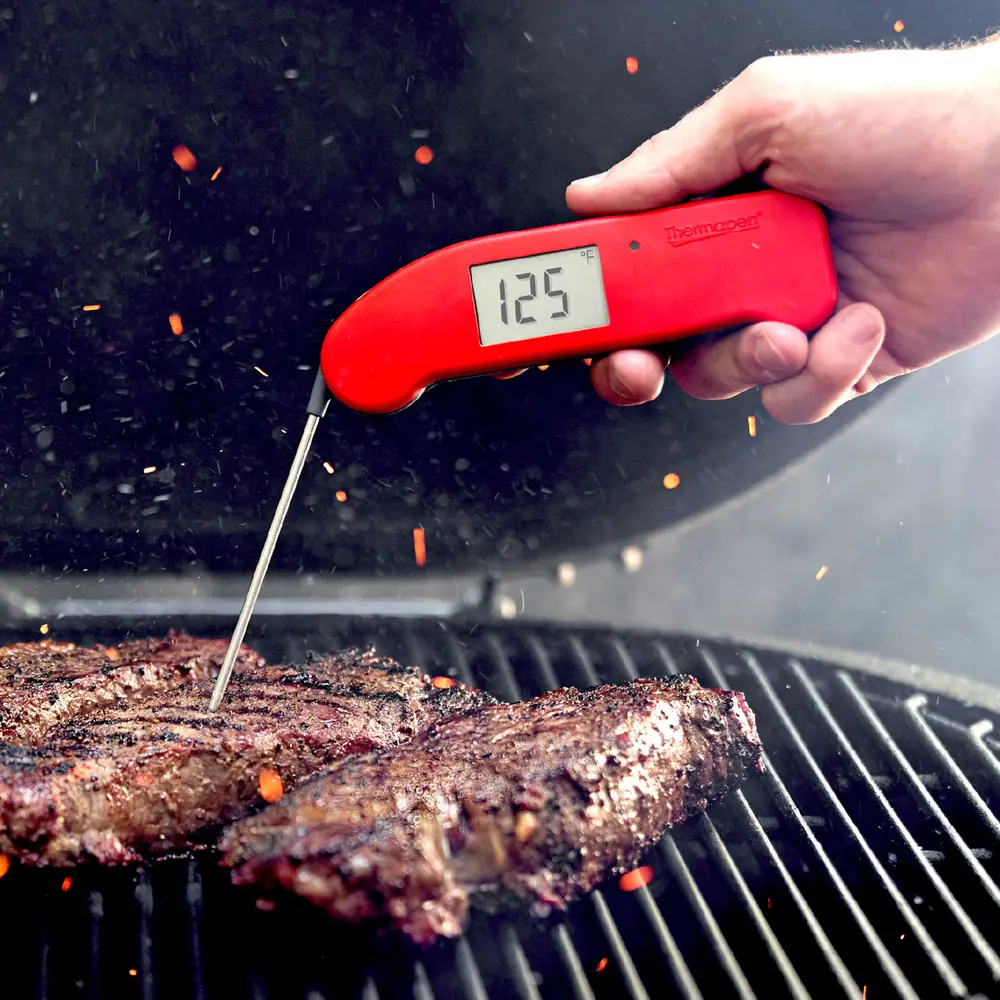 A hand holding a red Thermapen ONE reading 125 degrees, probing a steak on a charcoal grill with embers flying past