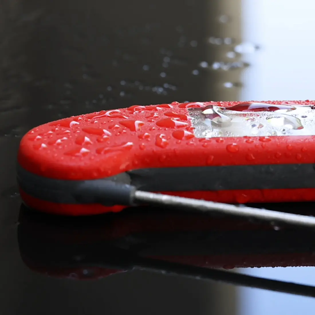 Close-up of a red Thermapen ONE lying on a wet dark surface, beaded with water droplets