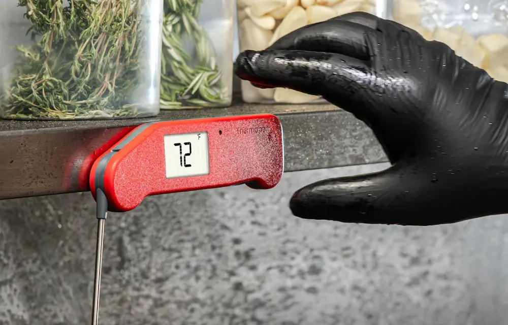 A red Thermapen NANO held by its integrated magnets against the vertical steel side of a prep counter, a gloved hand reaching for it
