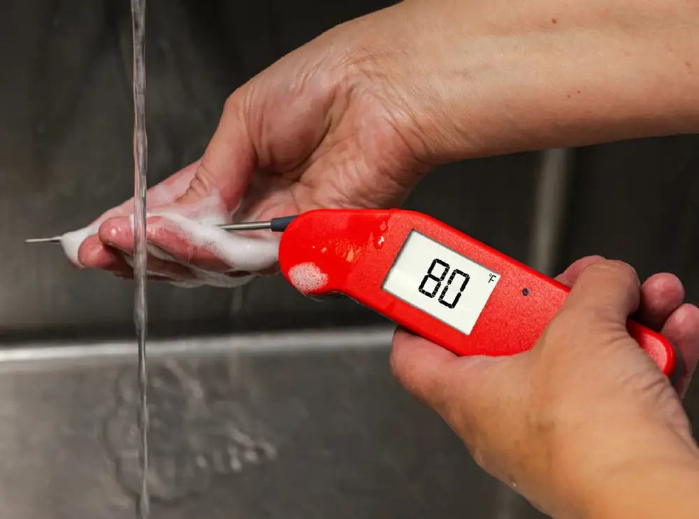 Two hands rinsing the steel probe of a red Thermapen under running water at a sink, the display reading 80