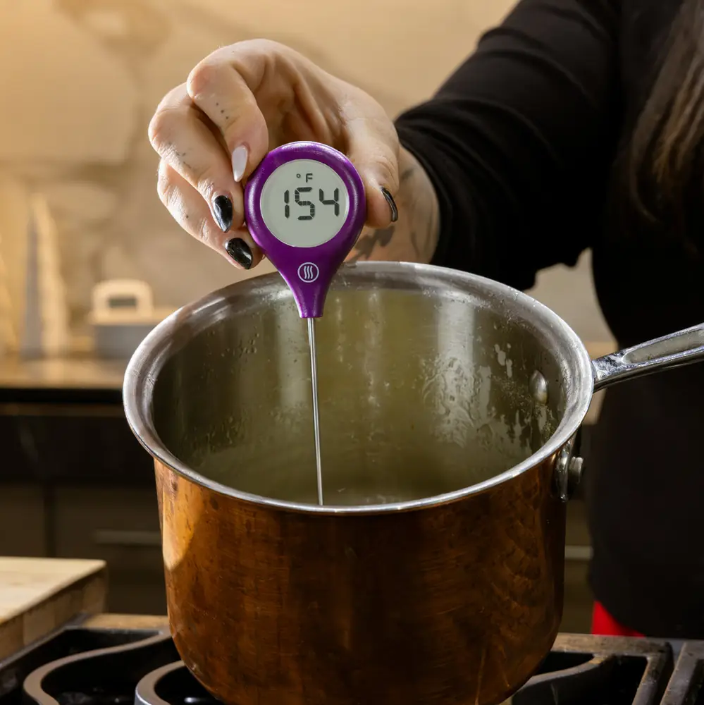 A hand holding a purple ThermoPop thermometer with the probe submerged in a saucepan of hot oil on a gas range, reading 154 degrees