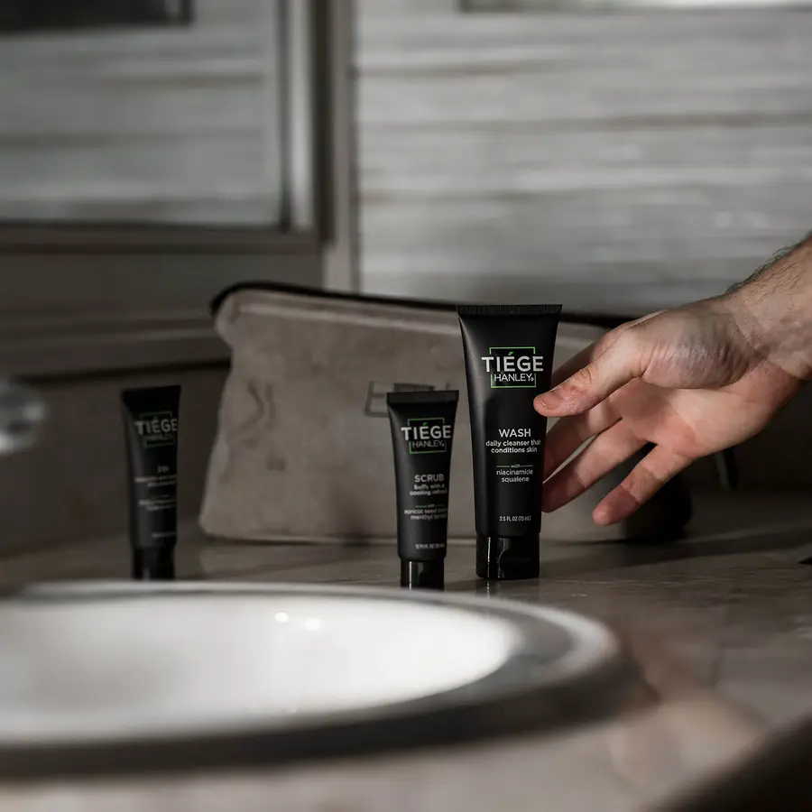 Tiege Hanley WASH, SCRUB and PM lined up on a bathroom counter next to a wash bag