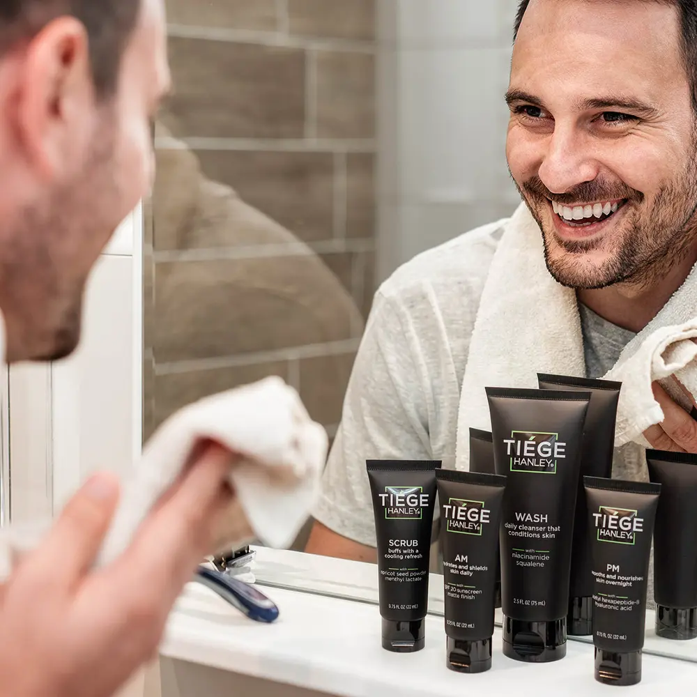 A man in a bathroom mirror with the Tiege Hanley system lined up on the counter