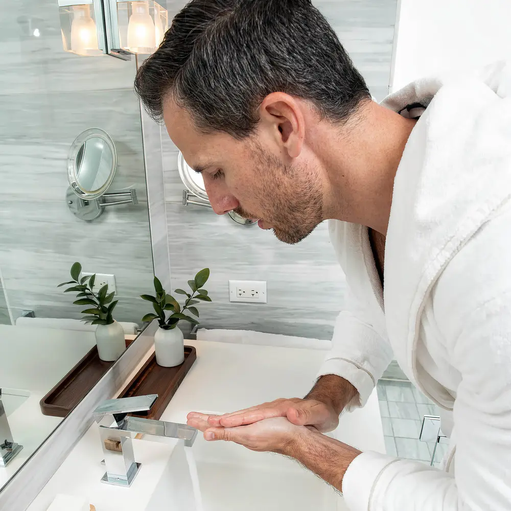 A man with grey at the temples washing his face at a bathroom sink