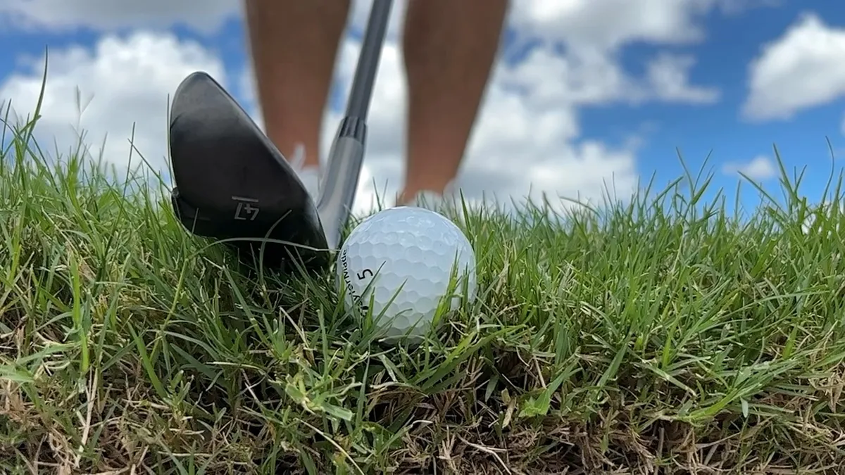 A wide soled black QX-1 wedge sitting behind a golf ball nestled down in thick rough