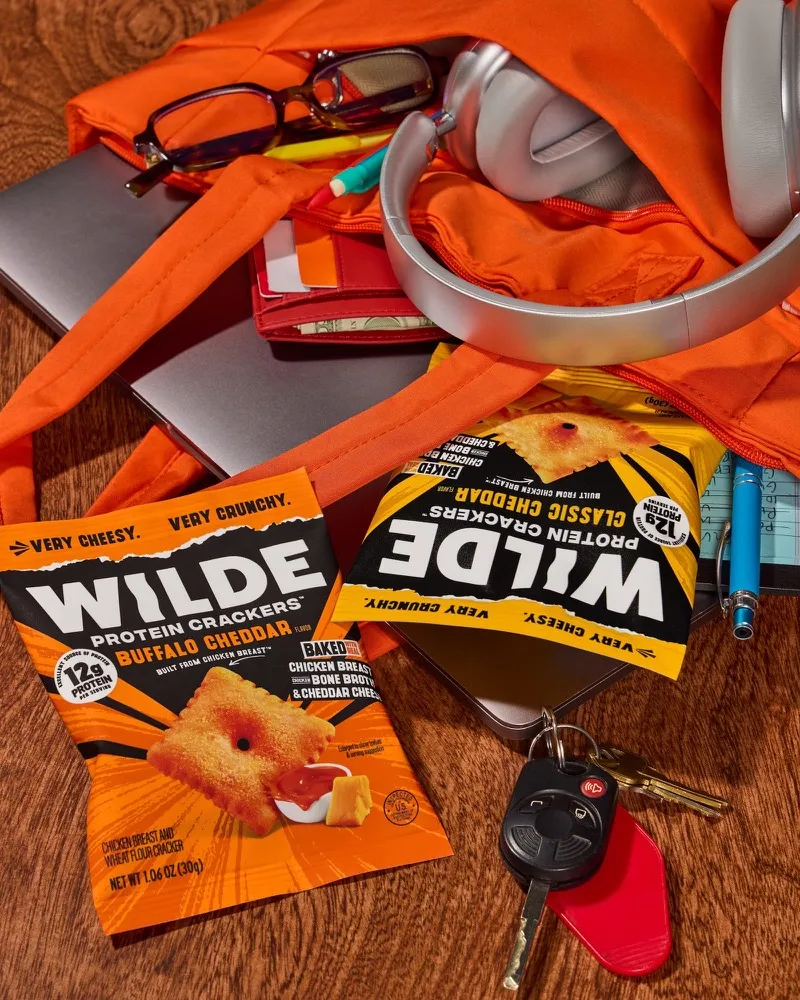 Two single serve WILDE cracker bags spilling out of a bag alongside keys, headphones and glasses