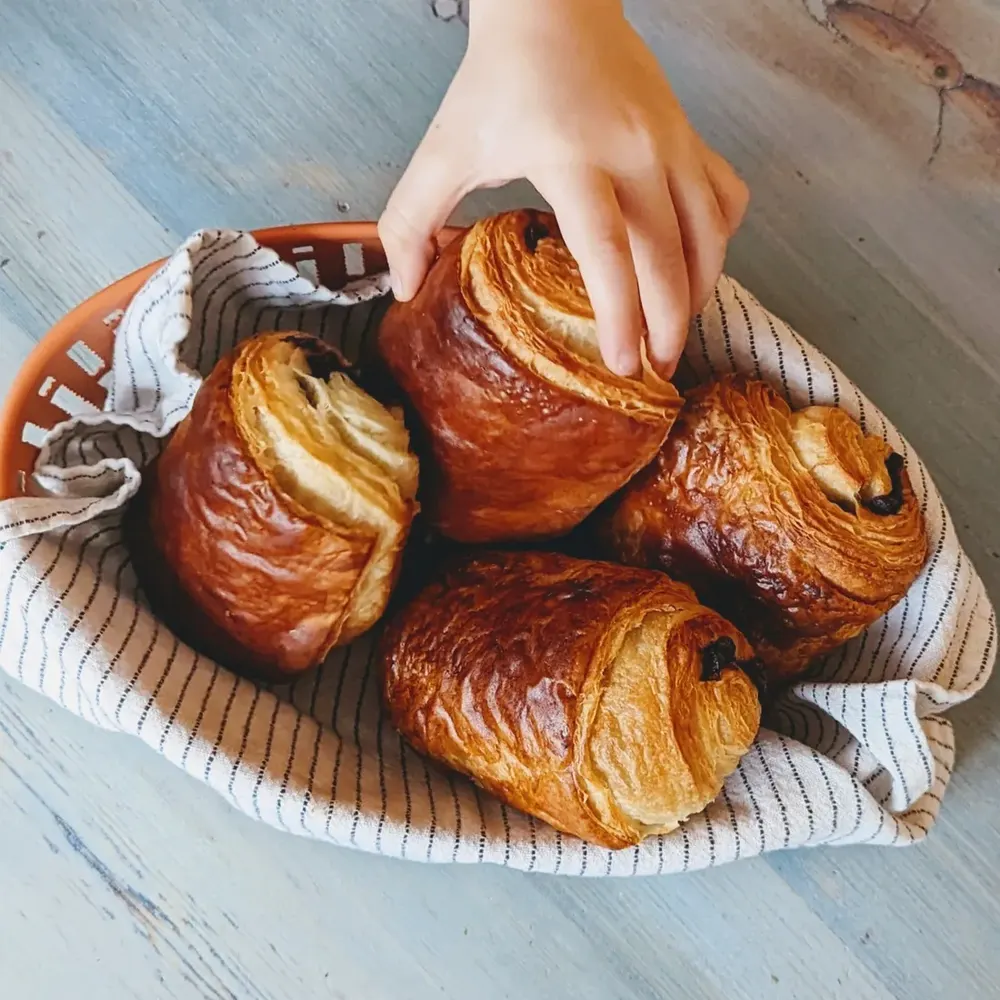 Close-up of dark golden pains au chocolat in a cloth-lined basket, the laminated layers clearly visible at the cut ends