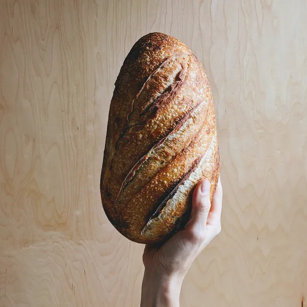 A hand holding a round scored sourdough loaf against a plain wooden wall