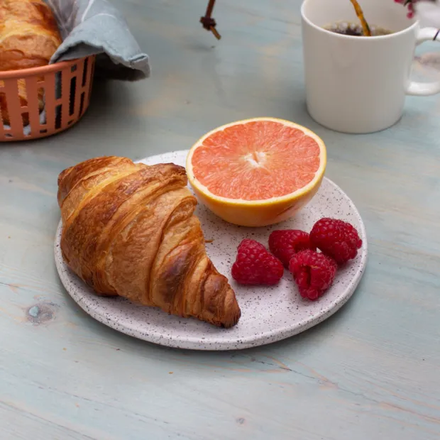 A croissant on a speckled plate with half a grapefruit and fresh raspberries, a mug of coffee behind