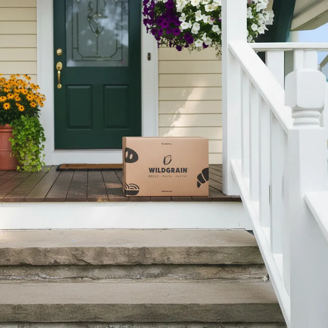 A Wildgrain cardboard delivery box on the steps of a house with a dark green front door and hanging flower baskets