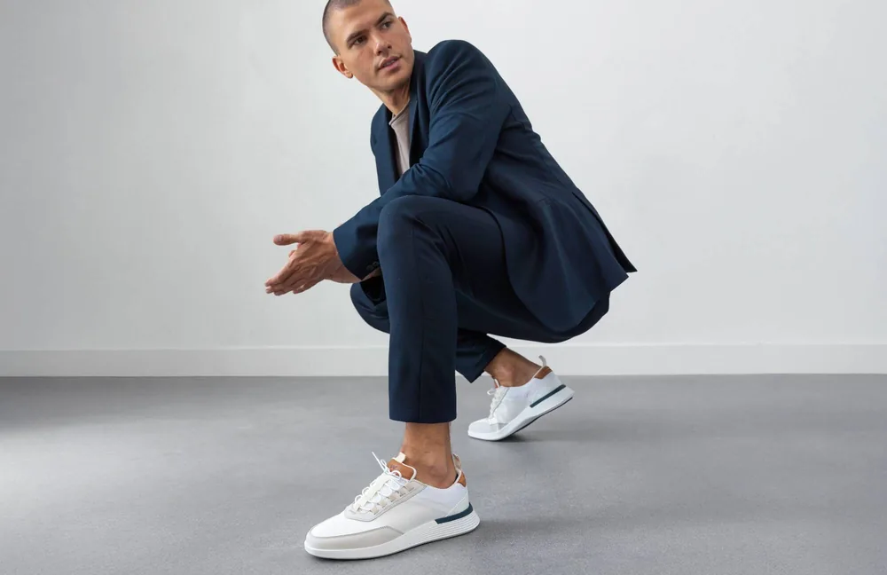 A man in a full navy suit crouching easily, wearing white Crossover Victory Trainers