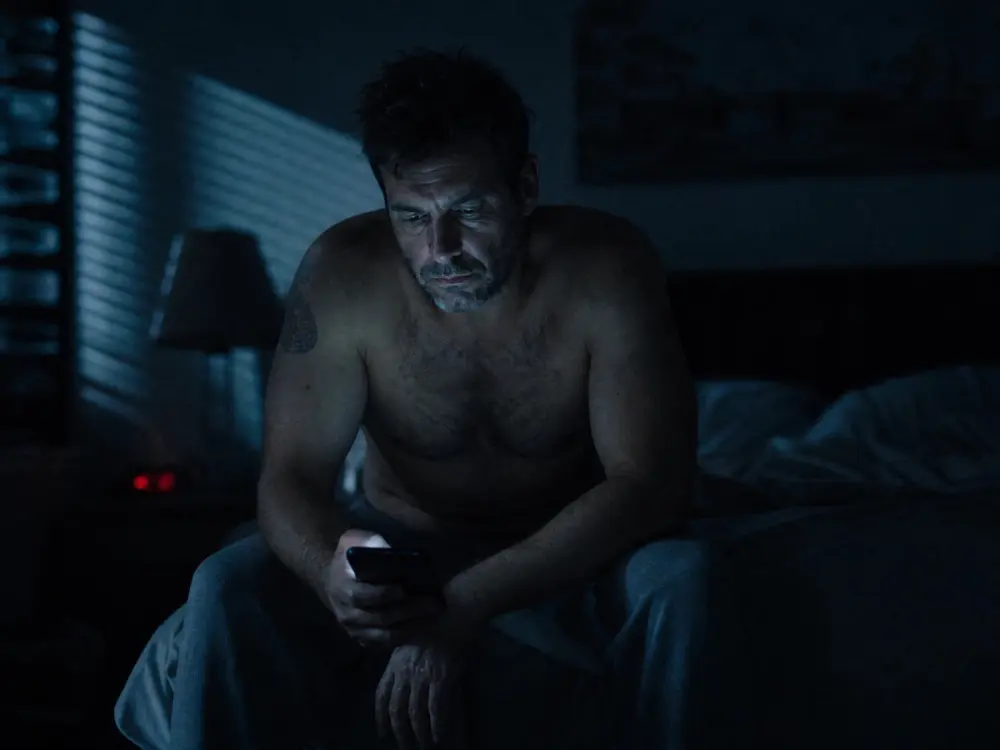 A man sitting awake on the edge of his bed at night, lit only by a phone screen